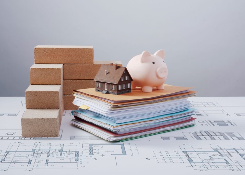 a piggy bank and miniature home sitting on top of a stack of paperwork and files, which is on top of a blueprint